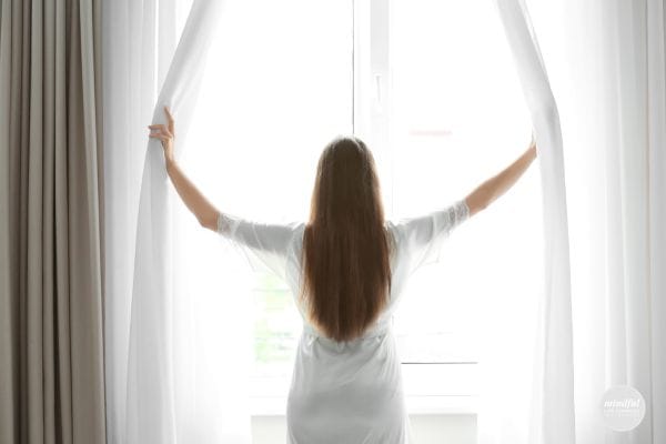 woman opening window shades to start the morning with positivity.